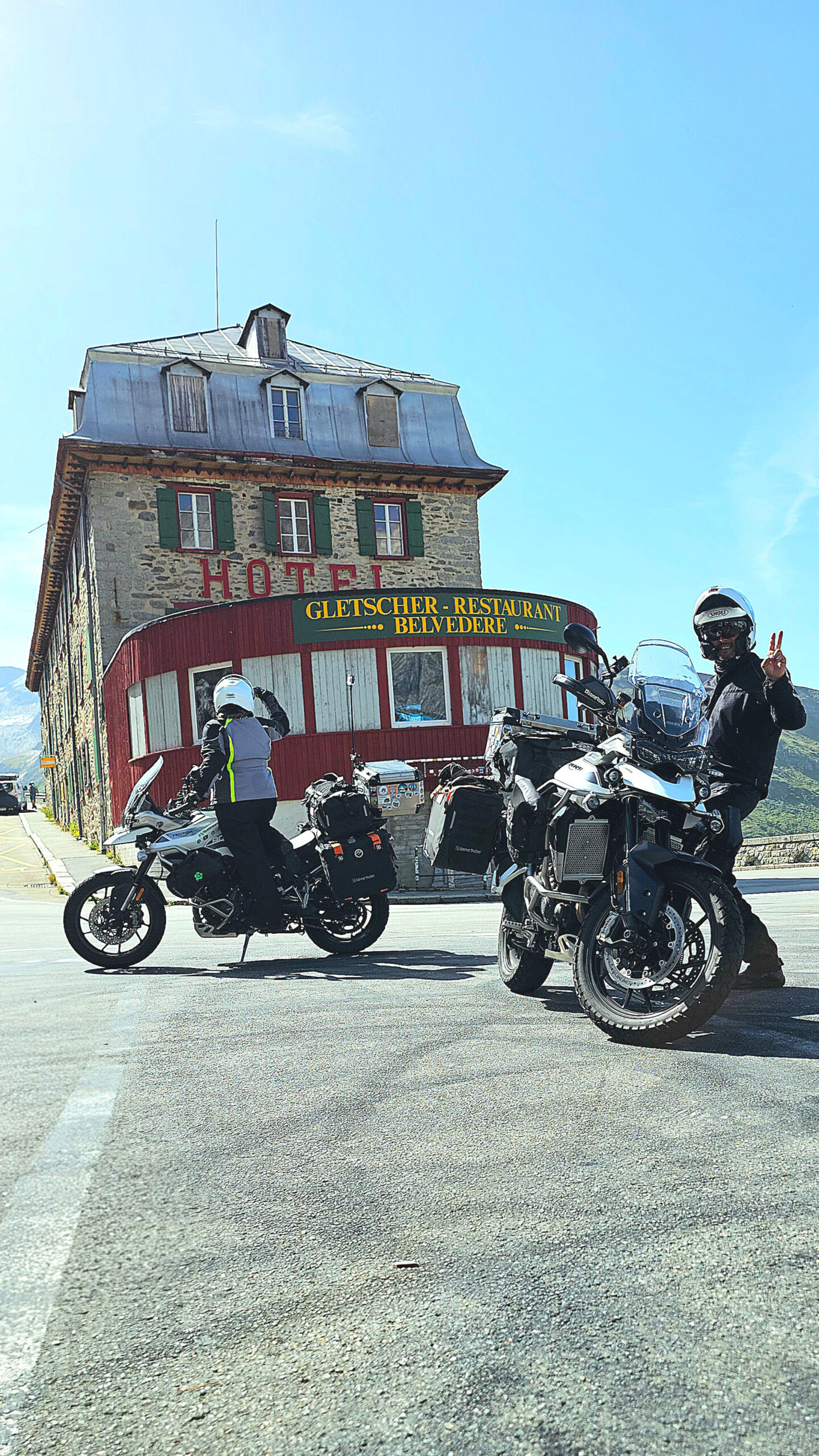 Motos Triumph Tiger garées devant l’hôtel Belvédère sur le Furka Pass, avec route alpine et glaciers en arrière-plan, ambiance James Bond