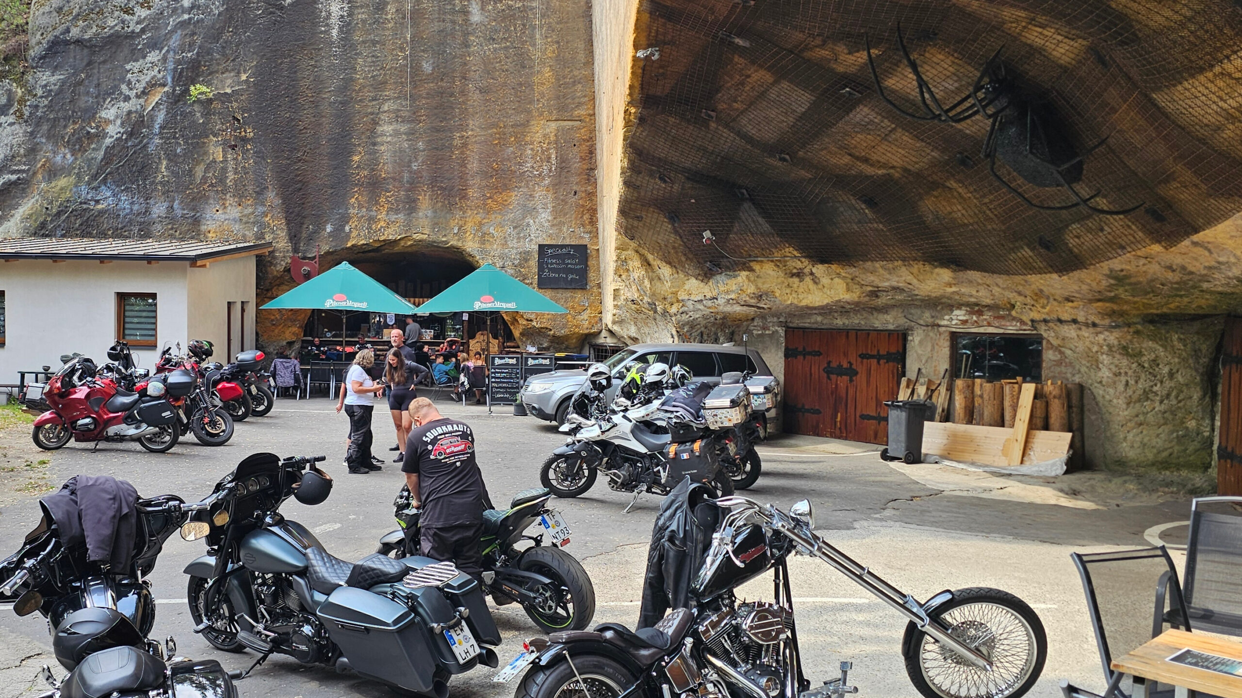 Photo prise devant la grotte en République tchèque : plusieurs motos stationnées, une gigantesque araignée en métal accrochée à la paroi rocheuse, et une conversation entre motards.