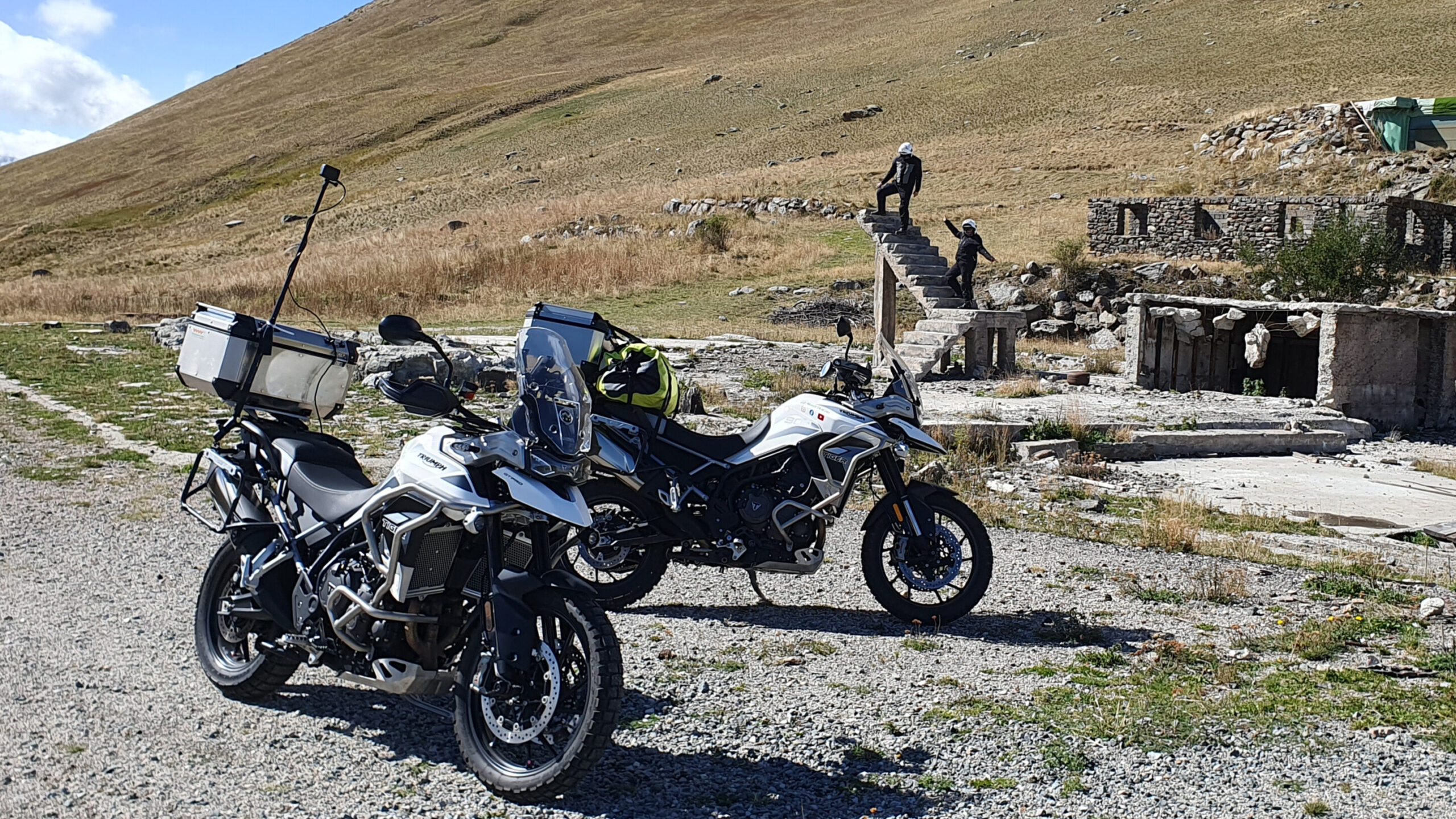 Motos arrêtées au sommet du col de Sarenne avec une perche à selfie GoPro visible au premier plan, et en arrière-plan un escalier en ruine isolé au milieu du paysage alpin.