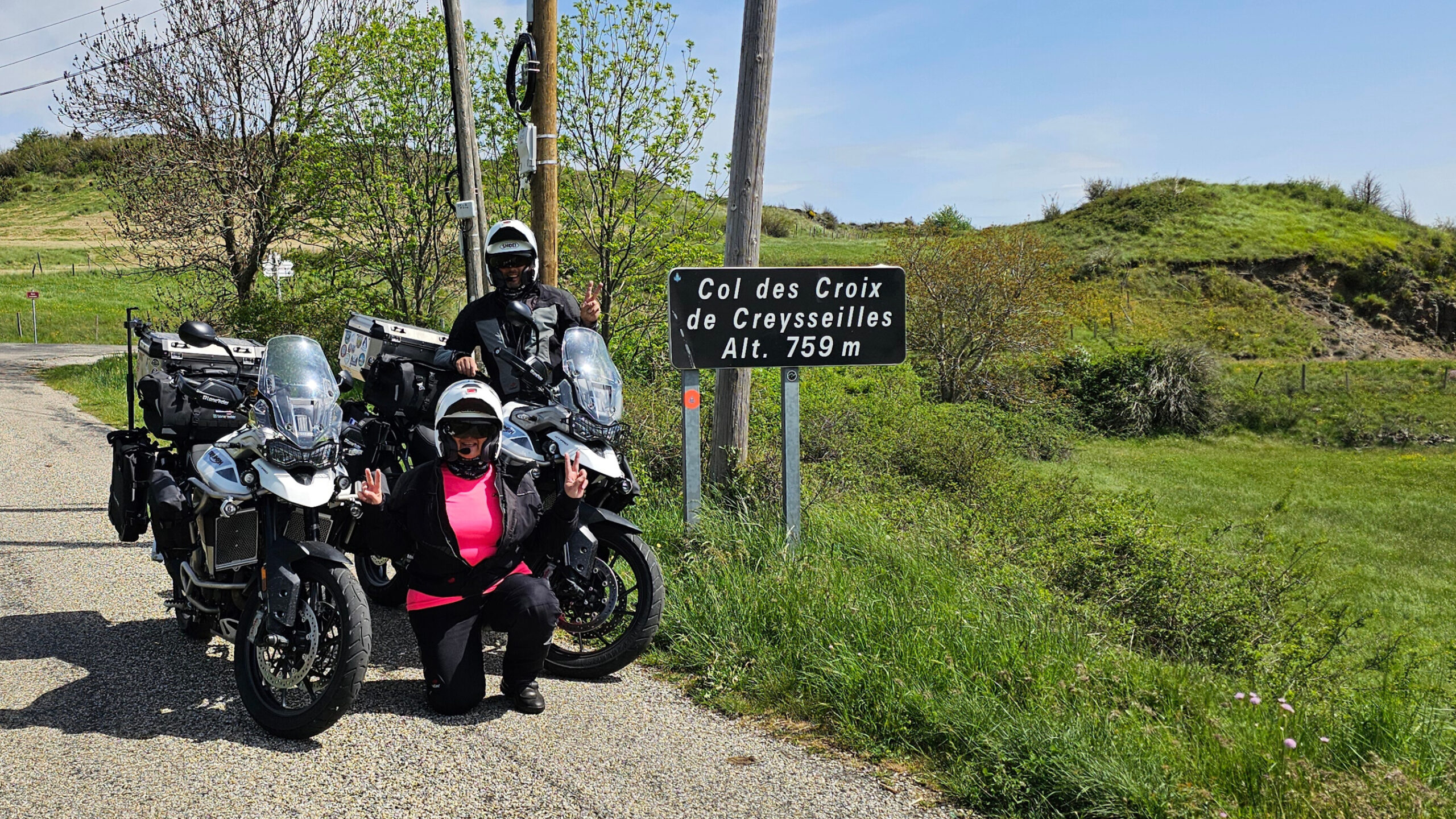 Deux motos Triumph Tiger 900 GT Pro arrêtées au col des Croix de Creysseilles, Cristian debout près du panneau et une motarde genou à terre entre les motos, équipées de sacoches Lone Rider.