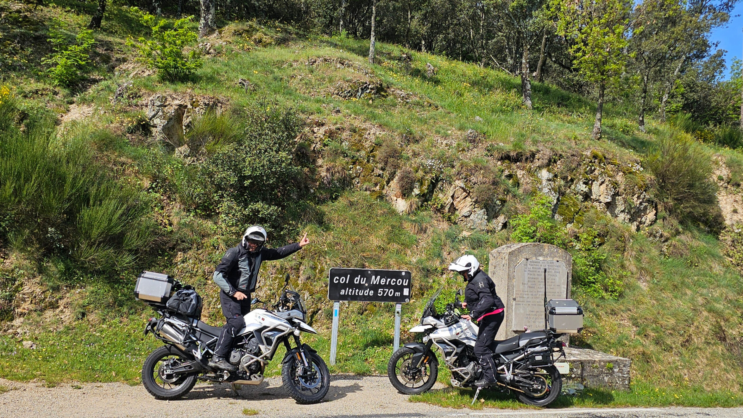 Deux motos Triumph Tiger 900 GT Pro stationnées à côté du panneau du Col du Mercou (570m), avec les pilotes équipés en tenues de moto Halvarssons.