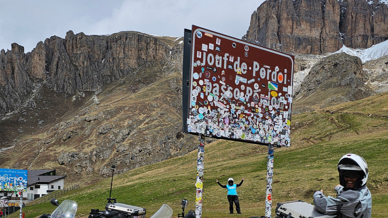 Sandrine au loin entre les poteaux du panneau officiel bilingue du Passo Pordoi et Cristian tout sourire au premier plan pointant ses bras vers elle, sur fond de crête montagneuse dans les Dolomites.