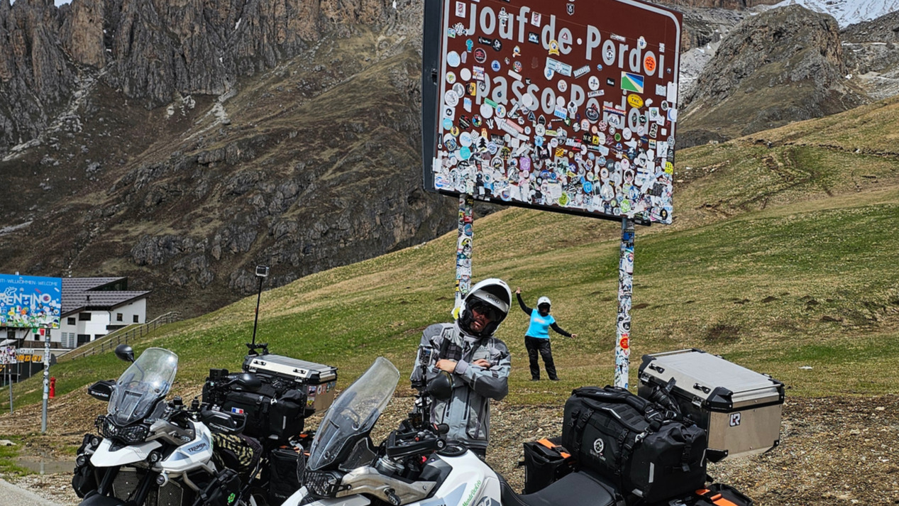 Les deux motos Triumph Tiger 900 GT Pro garées au Passo Pordoi près du panneau couvert d'autocollants, avec Cristian appuyé sur le poteau gauche et Sandrine mimant un avion au loin.