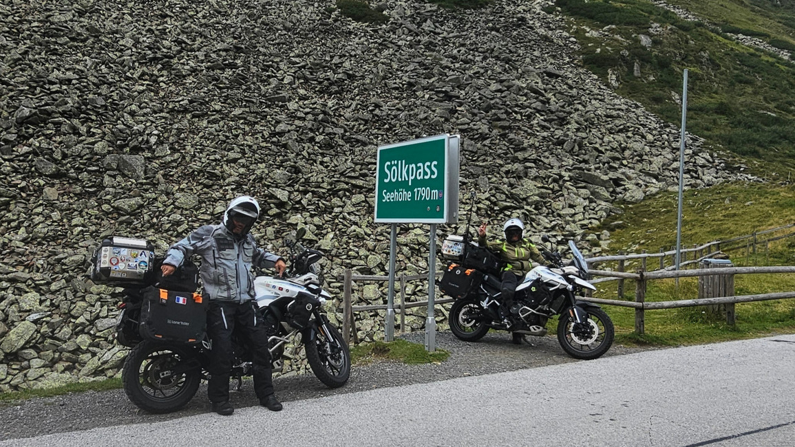 Gros plan de Sandrine et Cristian tout sourire à côté de leurs motos Triumph Tiger 900 au sommet du Sölkpass en Autriche.