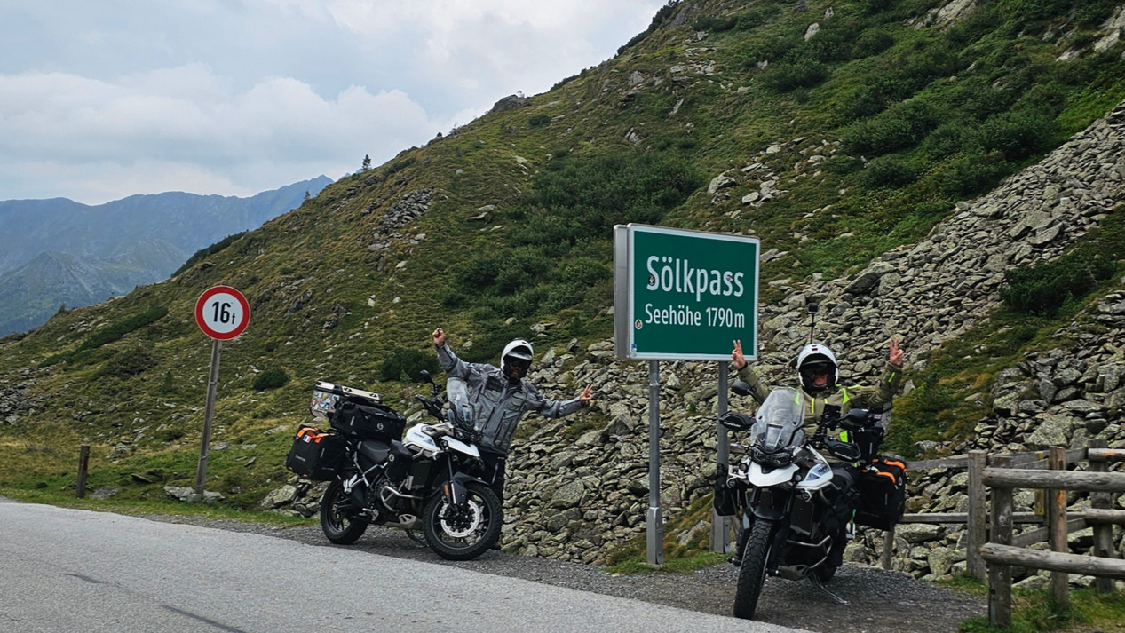 Deux motards en équipement Halvarssons faisant le signe V à côté de leurs motos Triumph Tiger 900 au sommet du Sölkpass, devant le panneau du col et l'interdiction aux plus de 16 tonnes.