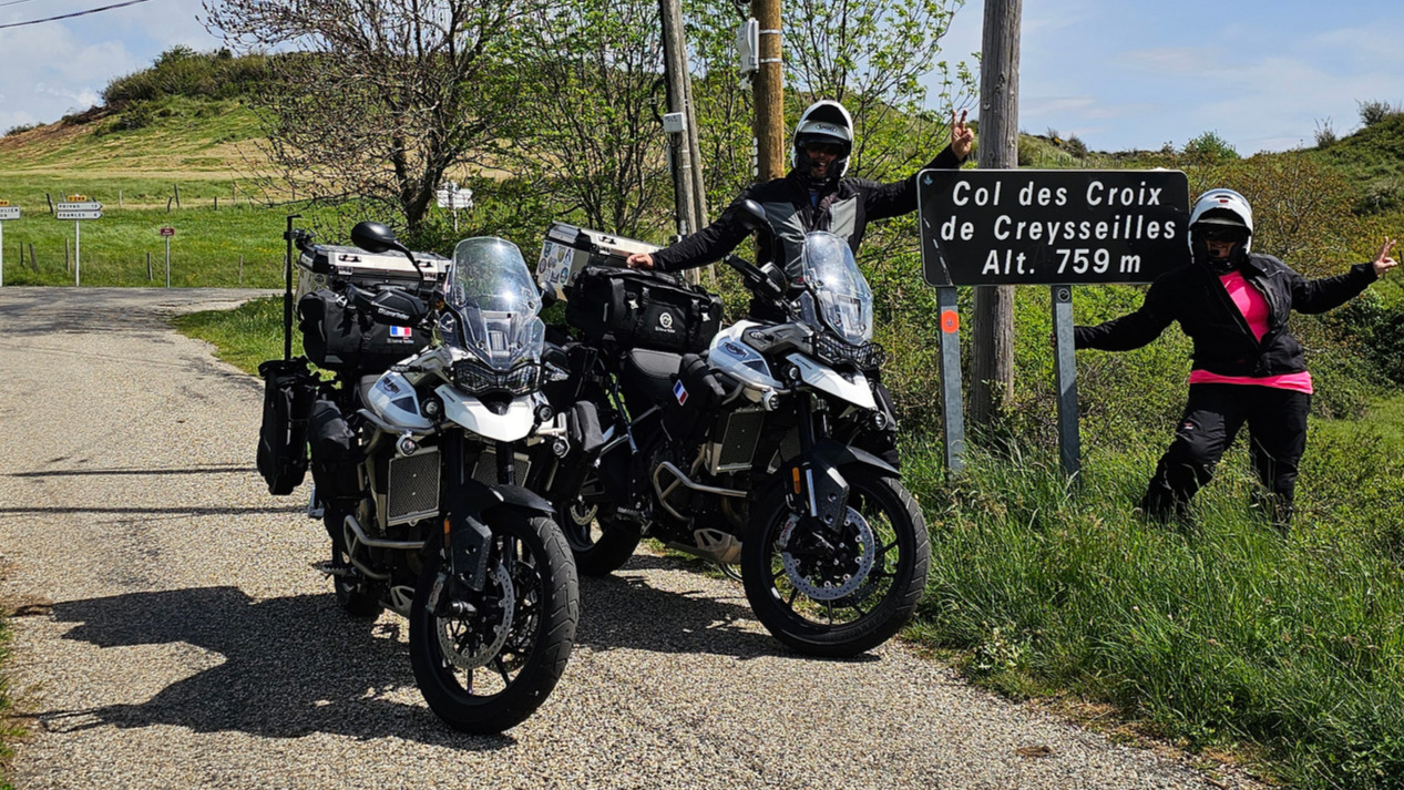 Motards faisant le signe V autour du panneau du col des Croix de Creysseilles sous un ciel bleu