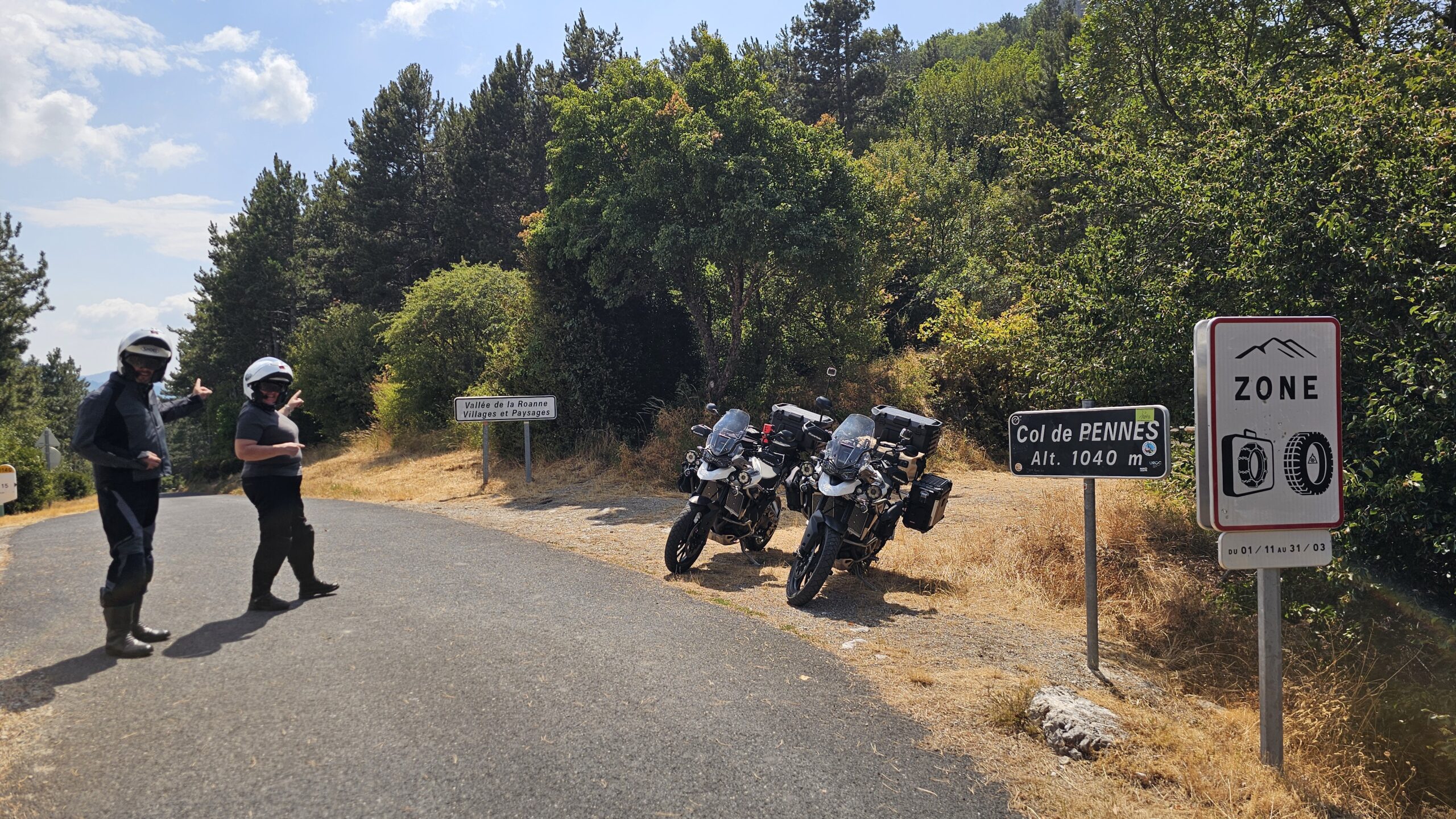 Pilotes en tenues Halvarssons au milieu de la route du Col de Penne devant le panneau de la vallée de Roanne