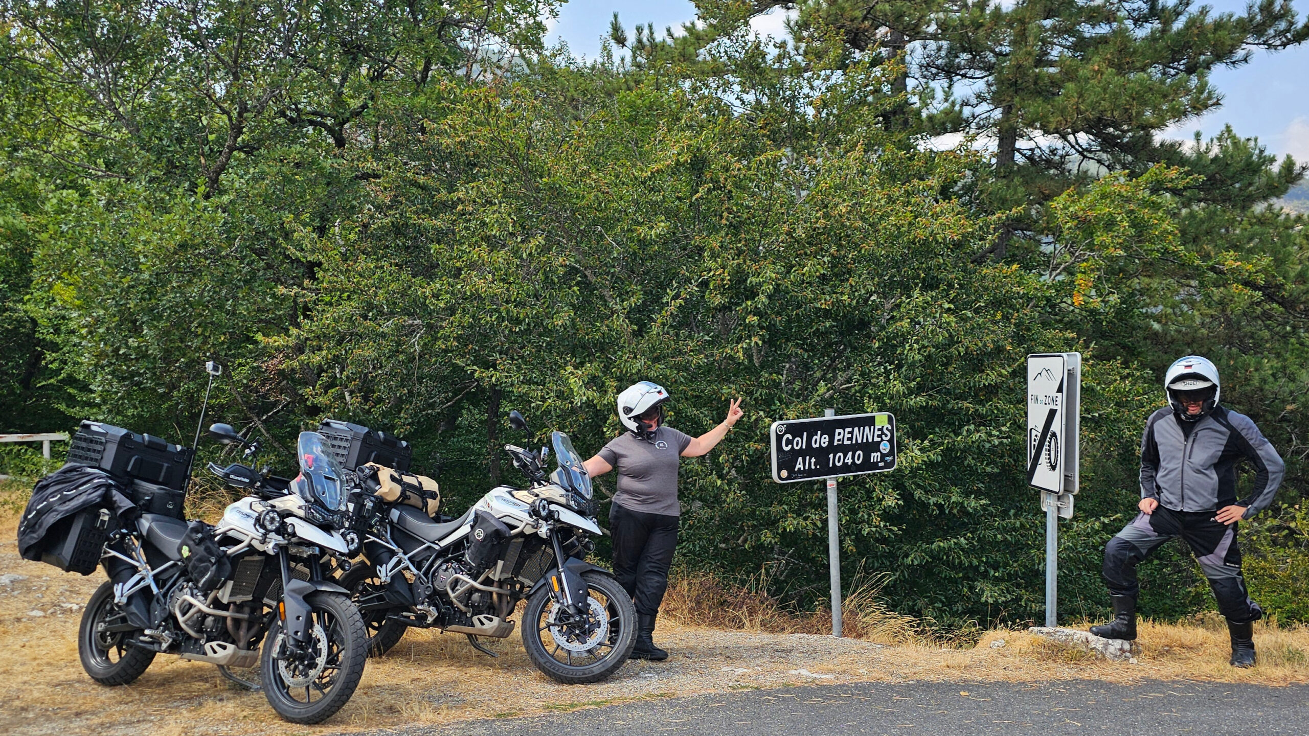 Motards pose autour du panneau du Col de Penne avec leurs motos garées à gauche