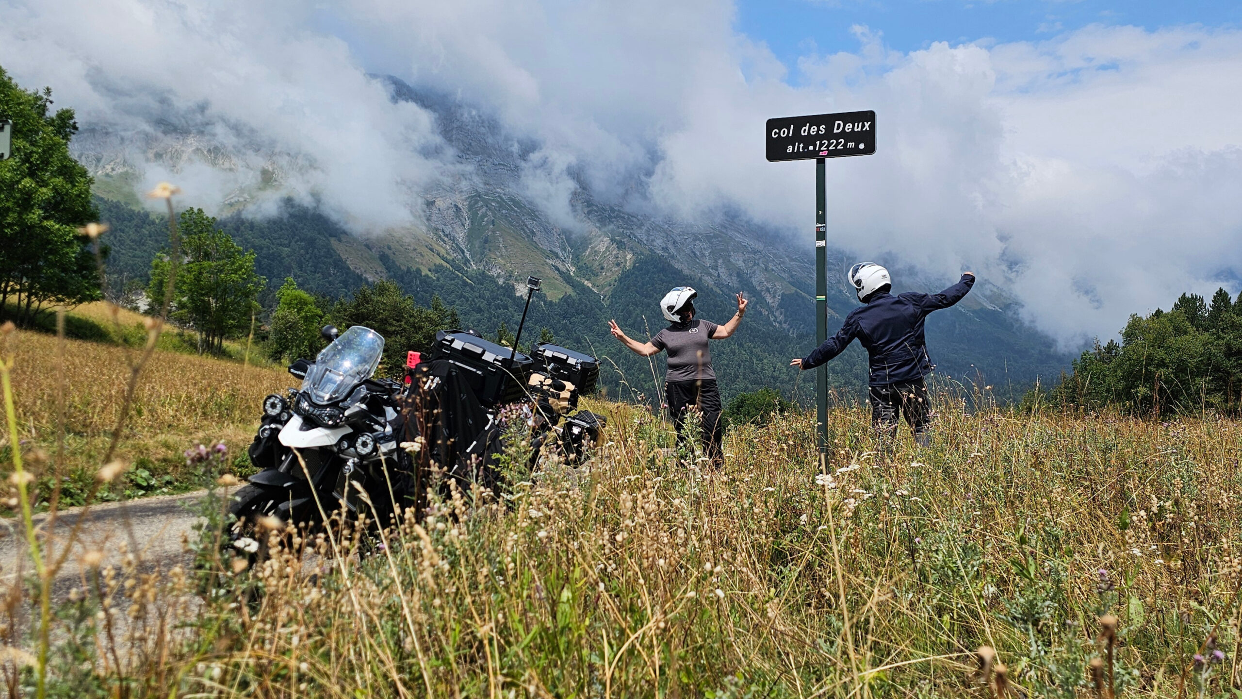 Pilotes en tenues Halvarssons et casques Shoei devant le panneau du Col des Deux avec deux motos Triumph Tiger 900 GT Pro bagagées en Lone Rider sous des nuages mystérieux.