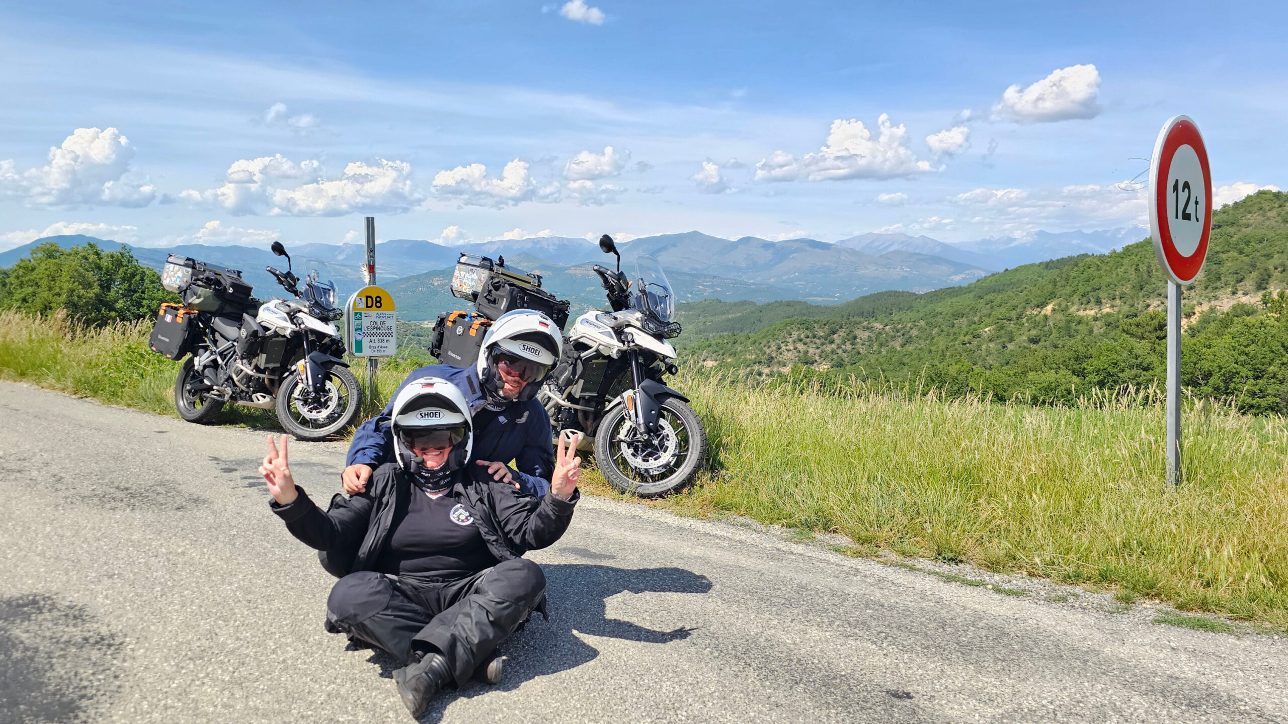 Motards assis au milieu de la route au Col de l'Espinouse, près de la borne à damier des cols remarquables et du panneau d'interdiction de plus de 12 tonnes.