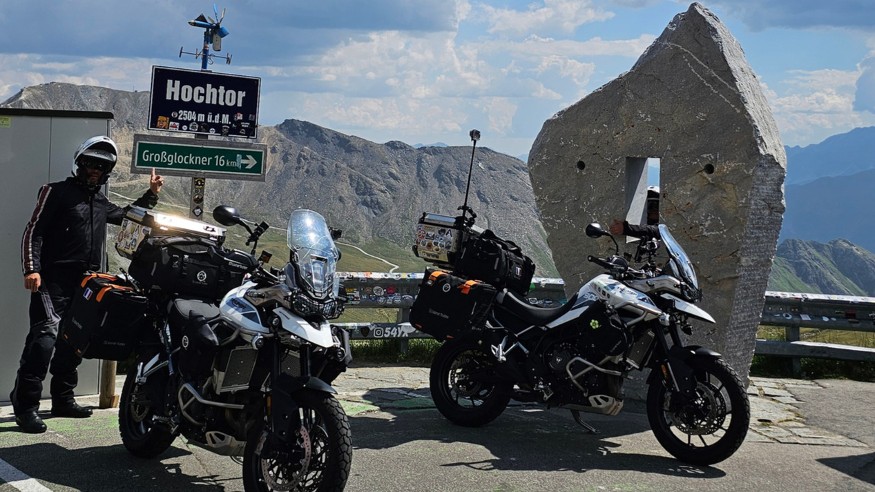 Cristian pose sous le panneau du Hochtor tandis que sa compagne apparaît à travers le trou du caillou percé.